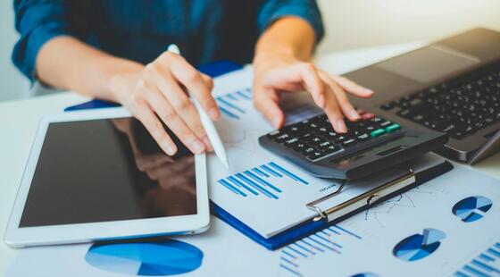 Persona manipulando una calculadora, un ordenador portatil, una tablet y algunos papeles con gráficos sobre una mesa de oficina.