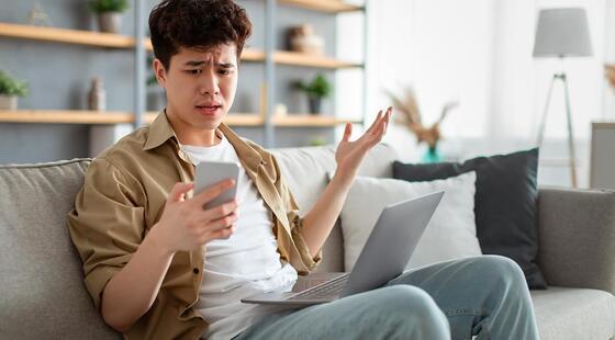 Hombre joven sentado en un sofá mirando con sorpresa e indignación un teléfono móvil