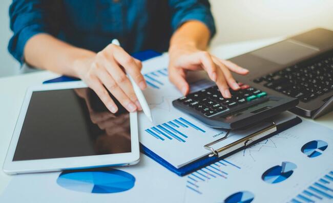 Persona manipulando una calculadora, un ordenador portatil, una tablet y algunos papeles con gráficos sobre una mesa de oficina.