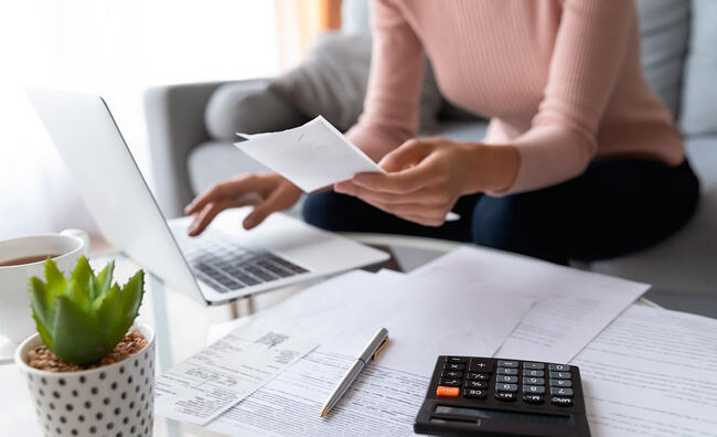 Mujer sentada enfrente de un ordenador portátil mientras sujeta un papel con una mano y teclea con la otra