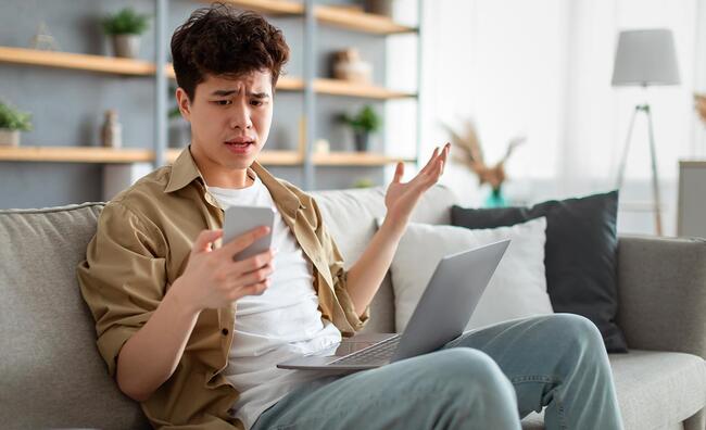 Hombre joven sentado en un sofá mirando con sorpresa e indignación un teléfono móvil