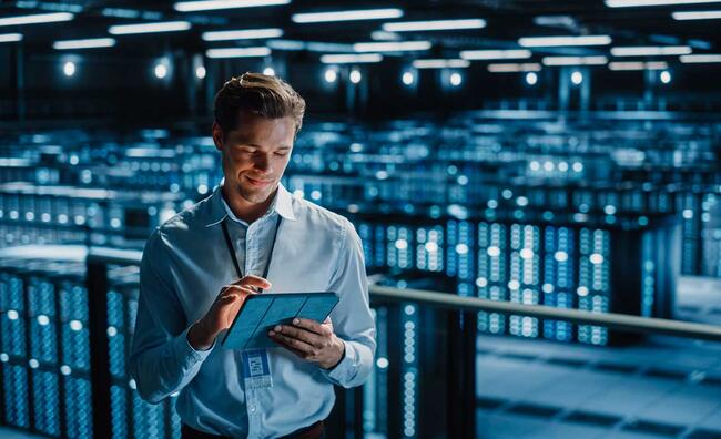 Hombre de pie manipulando una tableta electrónica en un espacio cerrado lleno de componentes electrónicos